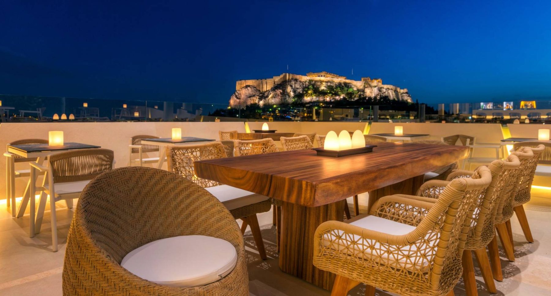 Rooftop dining table and chairs with the illuminated Acropolis in the background.