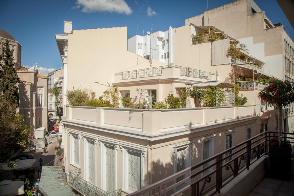 Corner building with pale upper floors and planted terraces beside a narrow Plaka street.