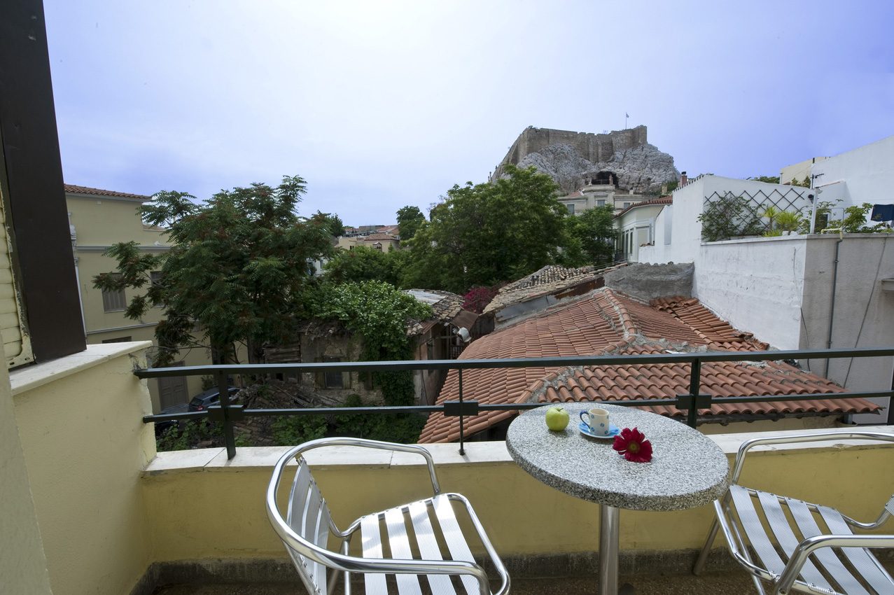 Small balcony with round table and two chairs overlooking tiled roofs towards the Acropolis.