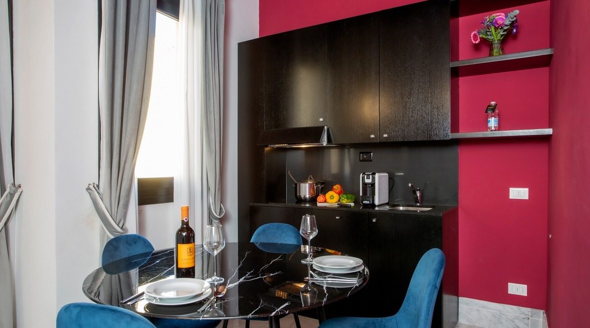 Compact dark kitchen beside a dining table with blue chairs.