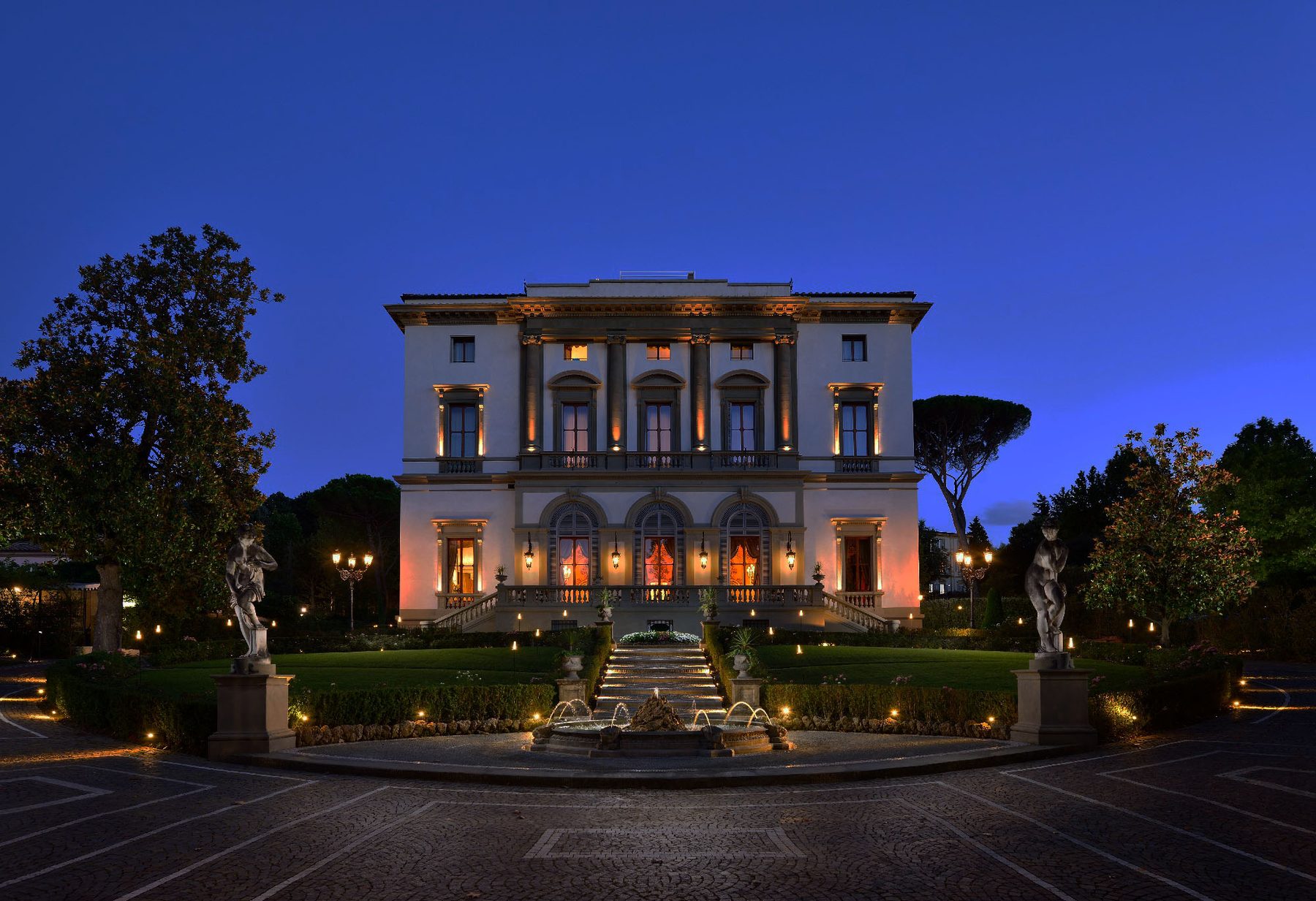 Illuminated villa facade behind garden paths, statues and a fountain.