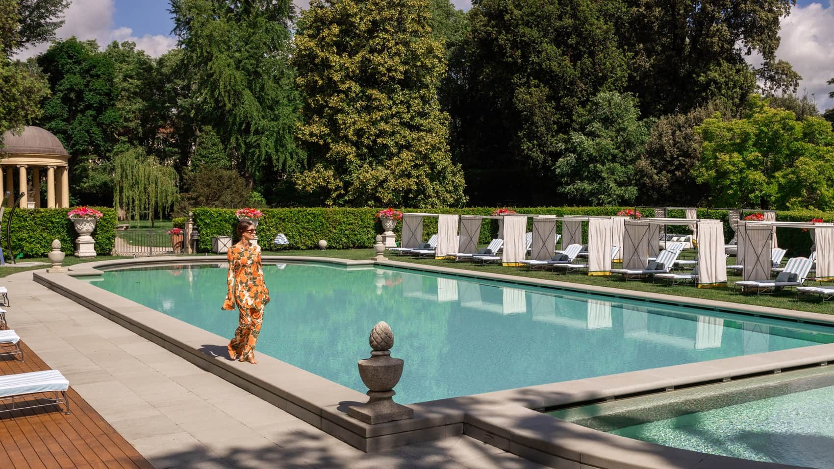 Outdoor swimming pool surrounded by trees, loungers and cabanas, with an adult beside the water.