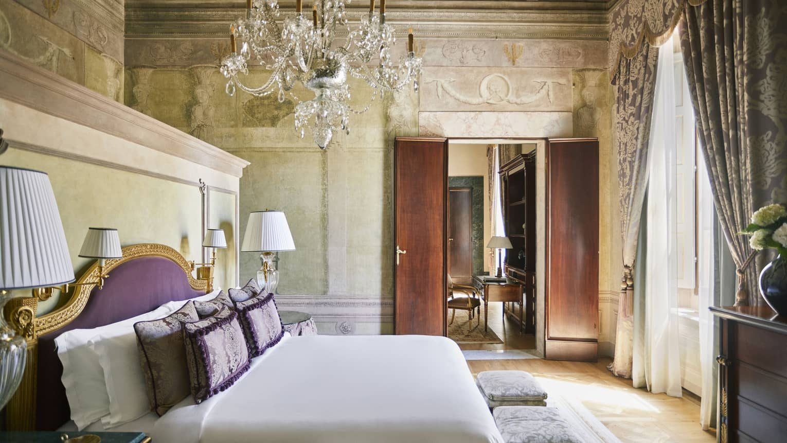 Bedroom with a purple headboard, chandelier and frescoed walls.