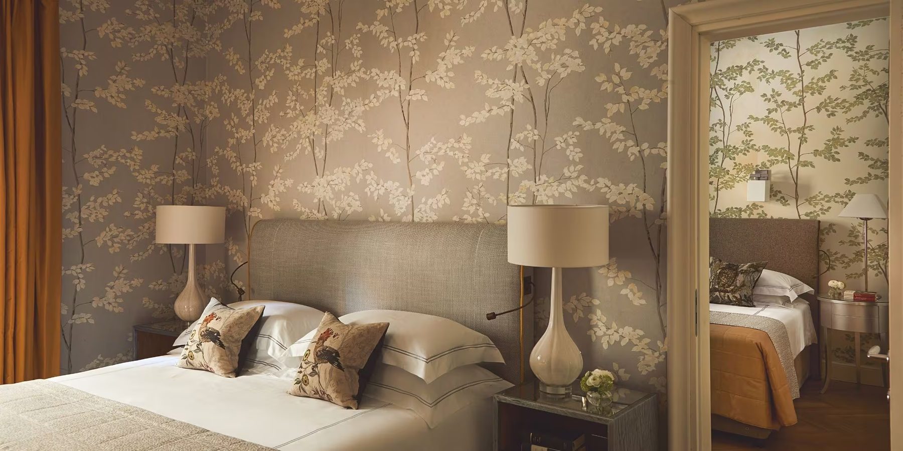 Bed with a padded headboard against botanical-patterned wallpaper.