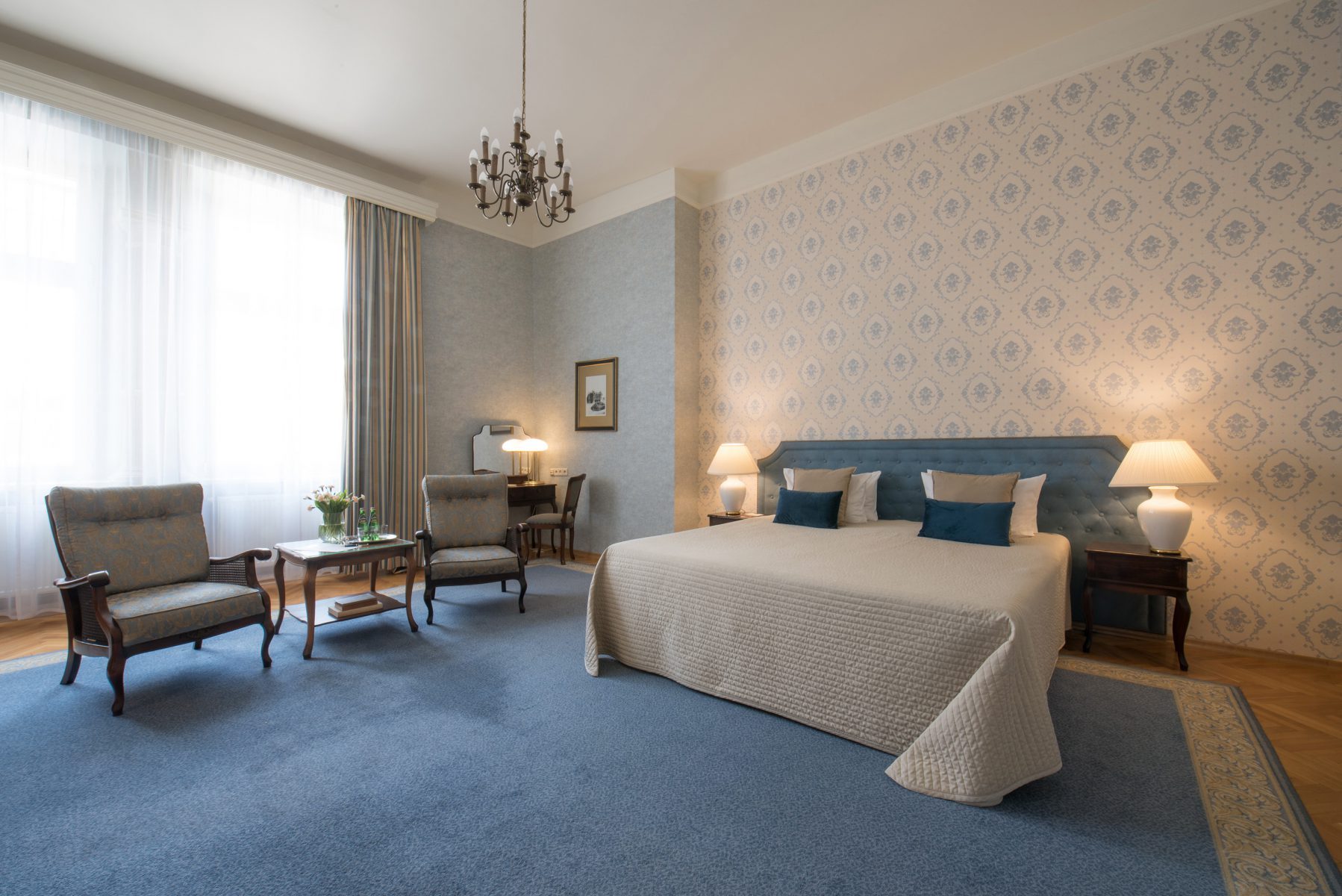 Grand Hotel Deluxe bedroom with blue accents and window-side chairs.