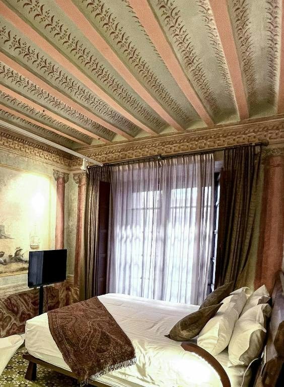Hotel bed beneath decorated ceiling beams beside a tall curtained window.