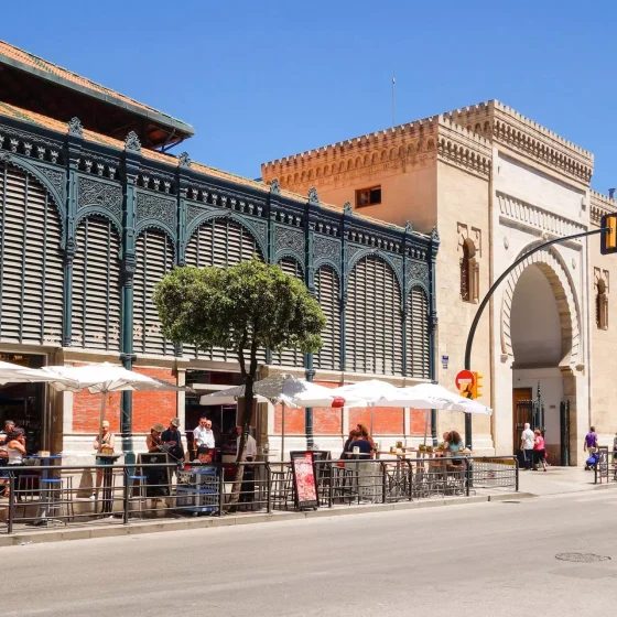 Atarazanas Market exterior with ironwork, a stone arch and pavement café tables.