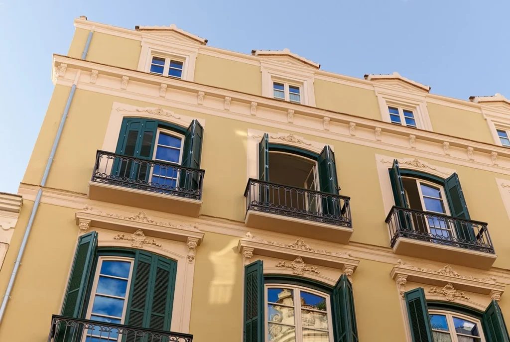Pale yellow historic façade with green shutters and small wrought-iron balconies.
