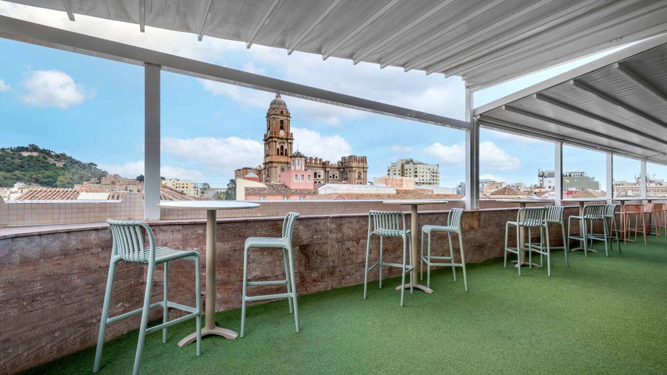 Rooftop bar stools beneath a canopy facing Málaga Cathedral and the Old Town skyline.