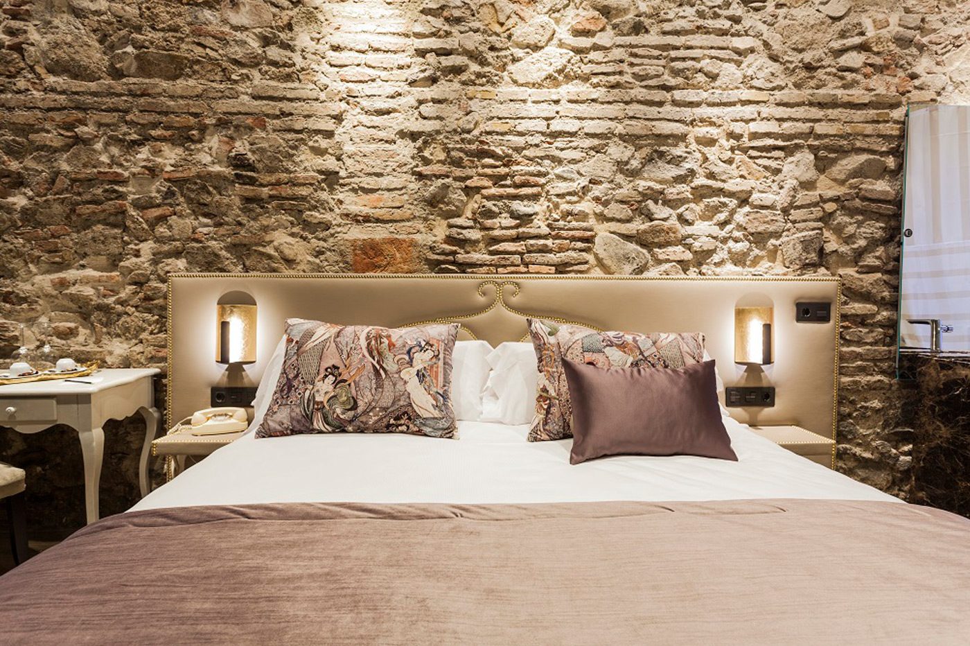 Bed and floral cushions beneath a softly lit exposed stone wall.