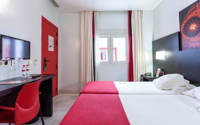 Twin beds with red throws, a red chair and black-and-red artwork in a hotel bedroom.