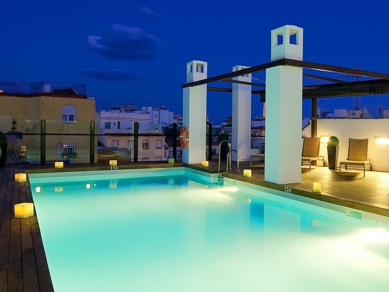Illuminated rooftop swimming pool with loungers and pergola structures at dusk.