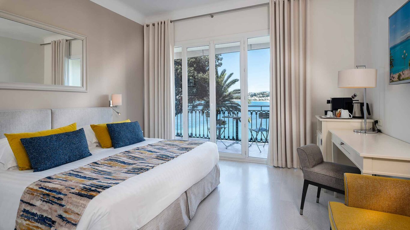 Bed and desk beside balcony doors overlooking the sea at Hôtel Suisse