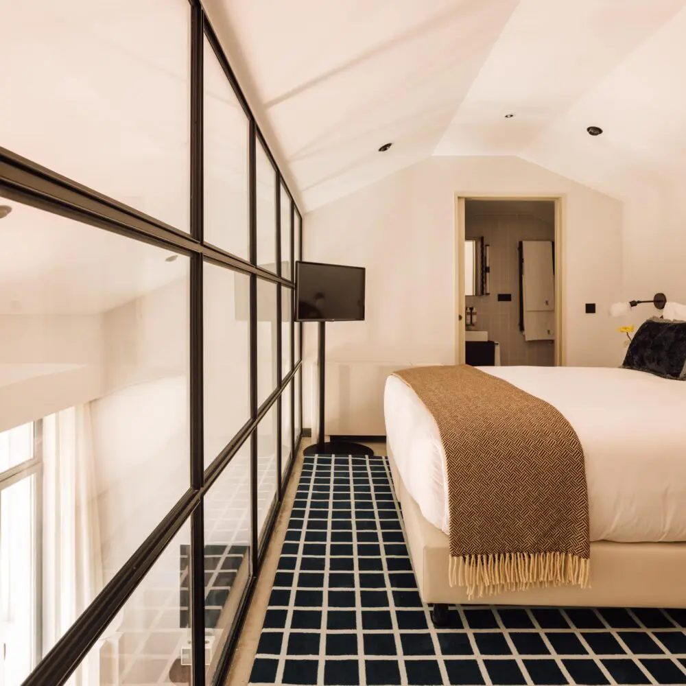 Bed, black-framed glass partition and checked rug in a mezzanine bedroom at The Rebello.