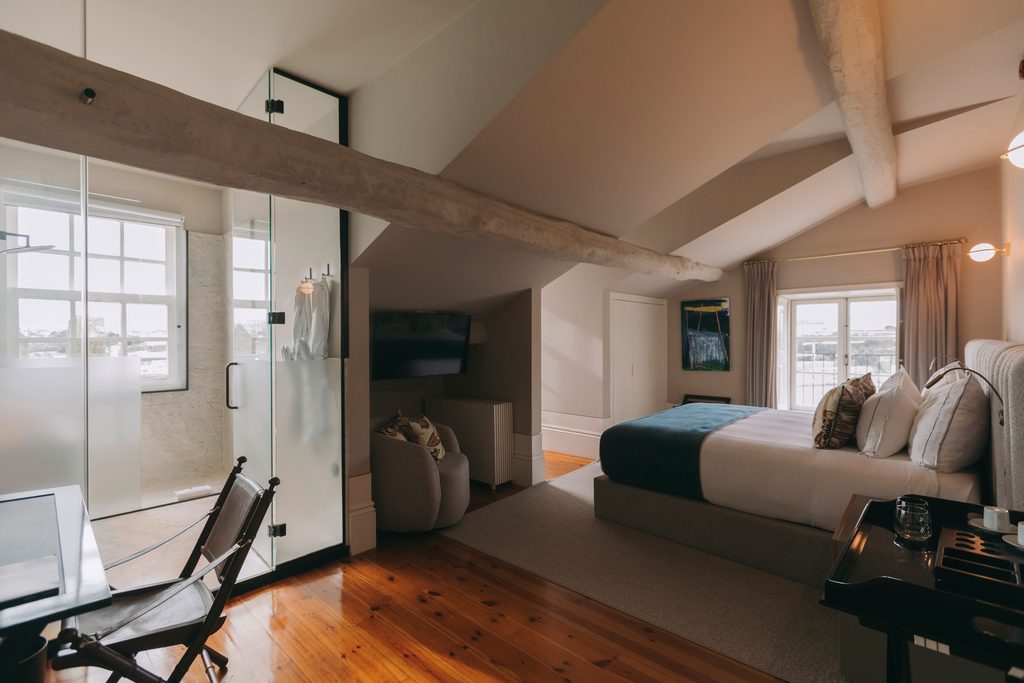Bed, timber floor and glazed shower partition beneath sloping beams at Torel Private Collection.