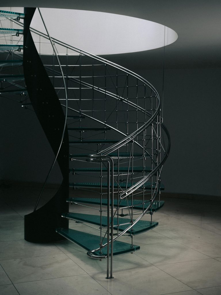 Curving glass staircase with a metal handrail below a circular opening.