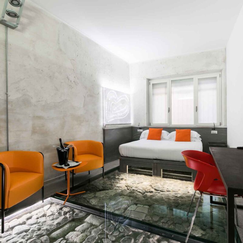 Bedroom with a white bed, orange chairs and a glass floor above exposed stonework.