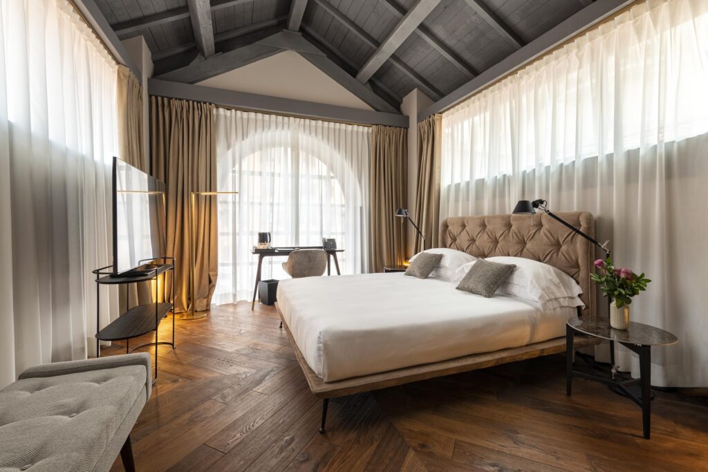 Bedroom beneath dark roof beams with tall curtained windows and a writing desk.