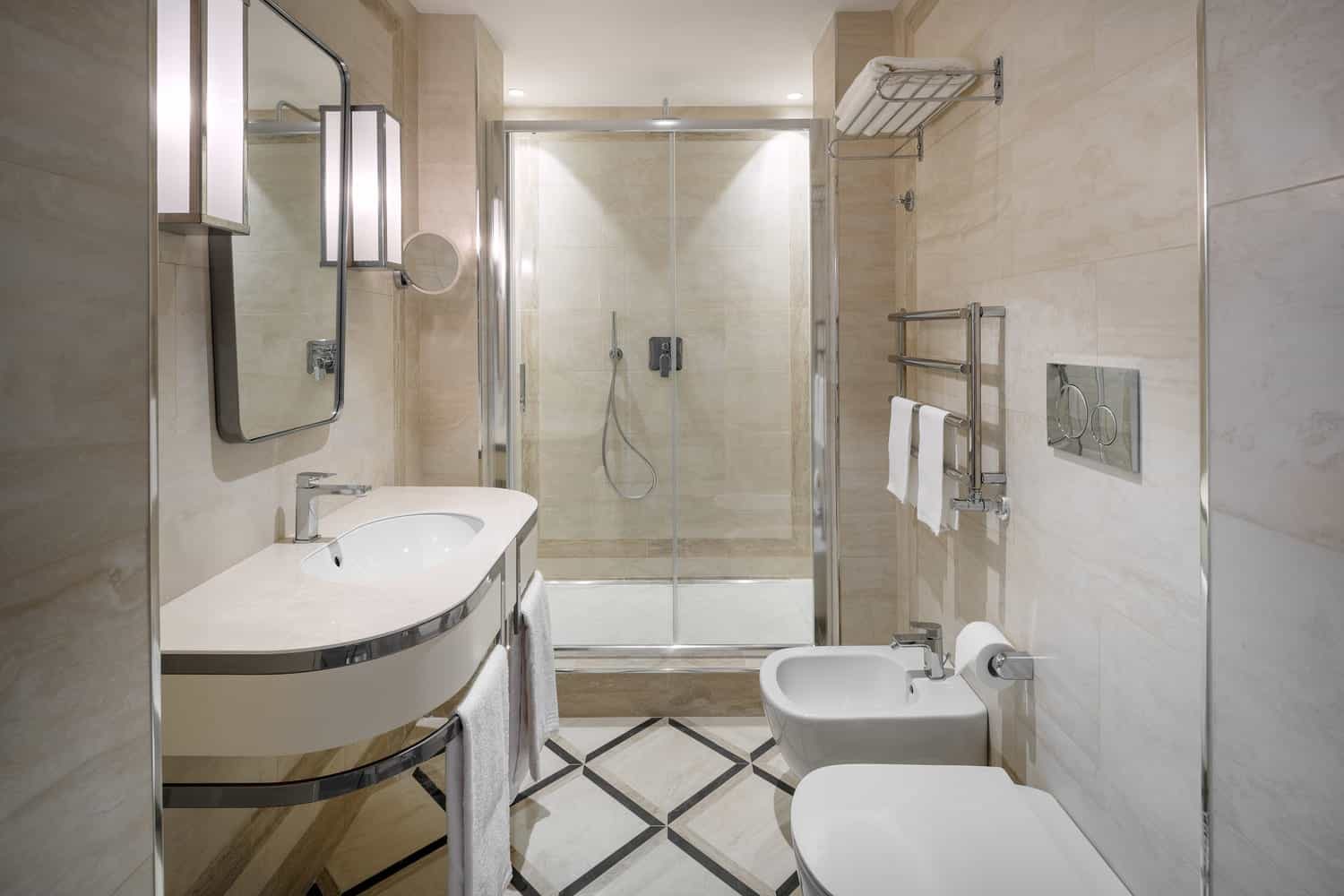 Glass shower enclosure beside a basin, toilet and bidet in a pale tiled bathroom.