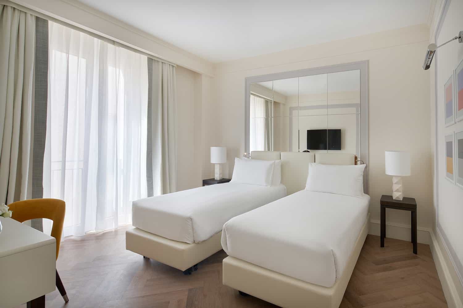 Two white single beds with bedside lamps and full-height curtains in a Deluxe room.