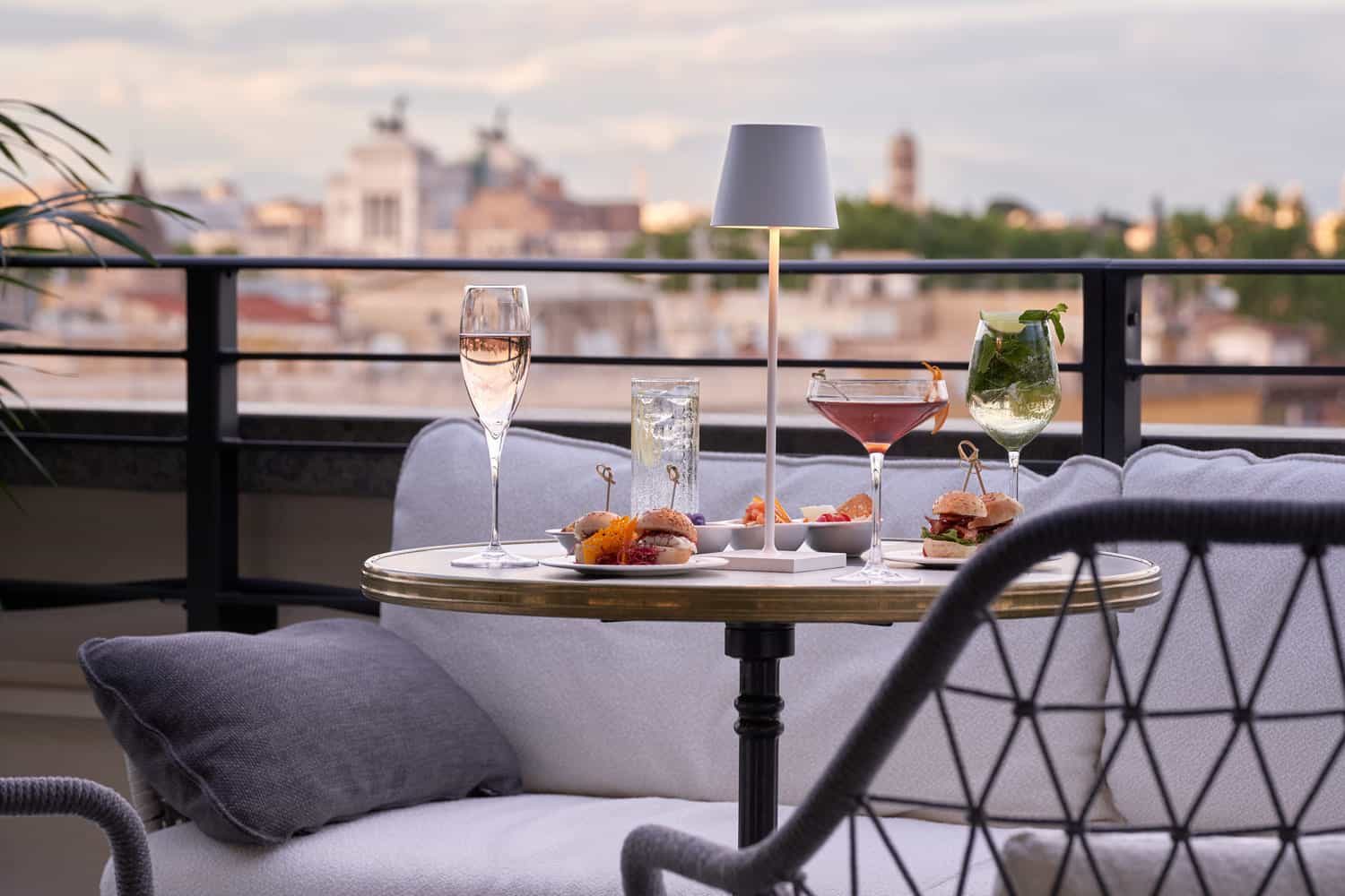 Outdoor table with glasses and small dishes beside cushioned seating and a view across rooftops.