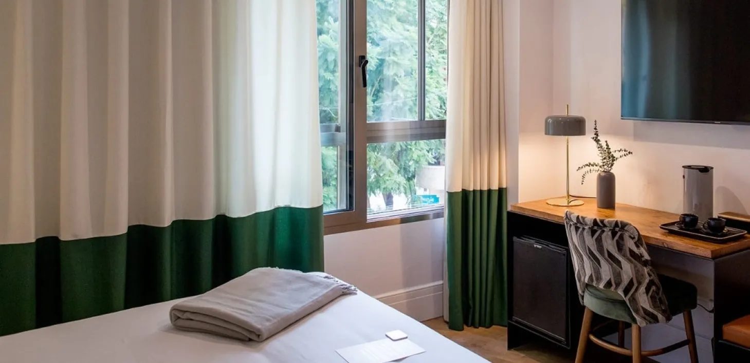 Foot of a bed, green-trimmed curtains, window and desk in Cosmo’s Superior Room category.