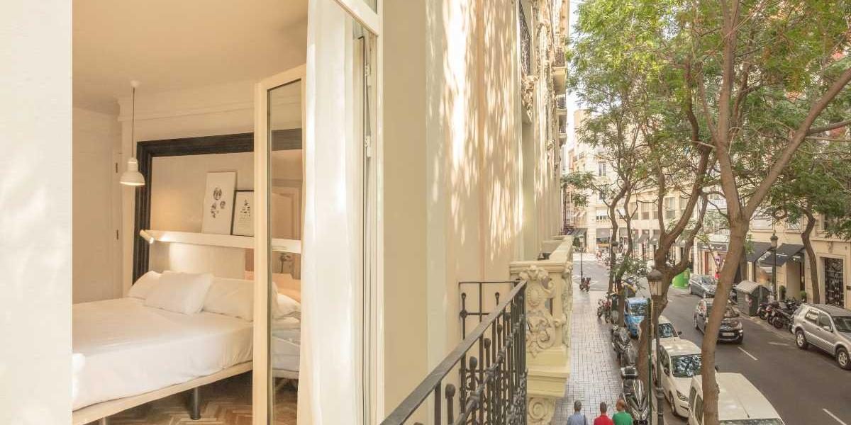 Wrought-iron balcony railing, trees and a street seen beside open bedroom doors.