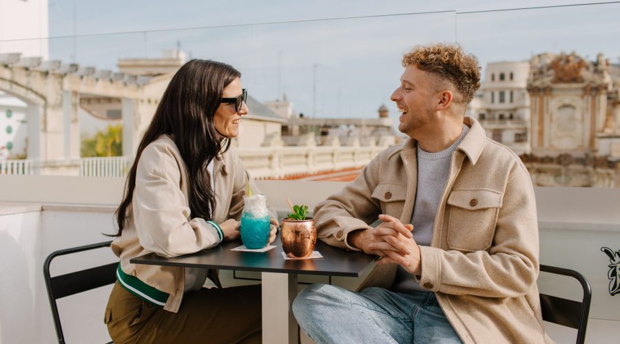 Two people seated with drinks on the Marqués House rooftop, with city buildings behind.