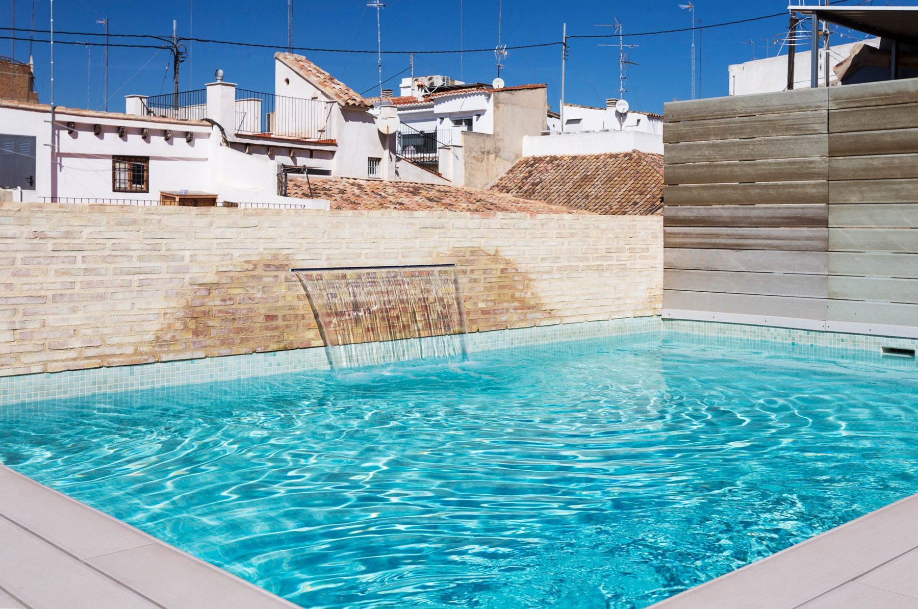 Blue rooftop pool with a wall-mounted water feature and city rooftops behind.