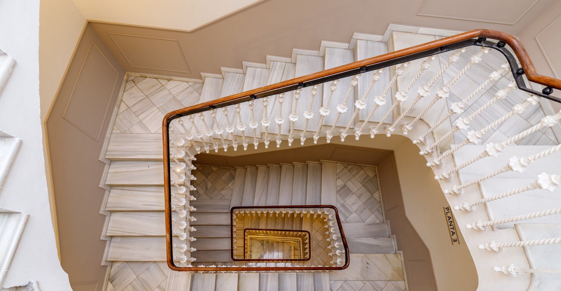 Looking down a square staircase with pale steps, decorative balustrades and timber handrails.