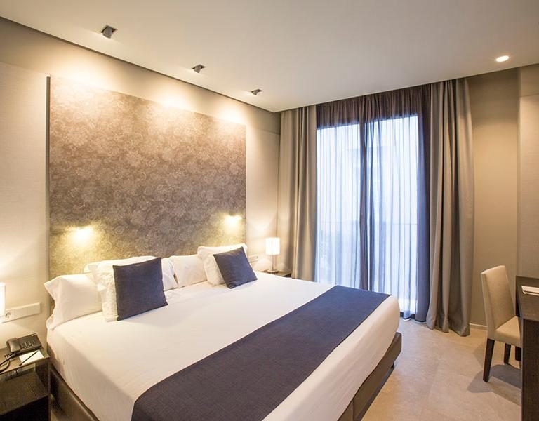 Bed with a blue runner, textured wall, curtains and desk corner in a Vincci Room.