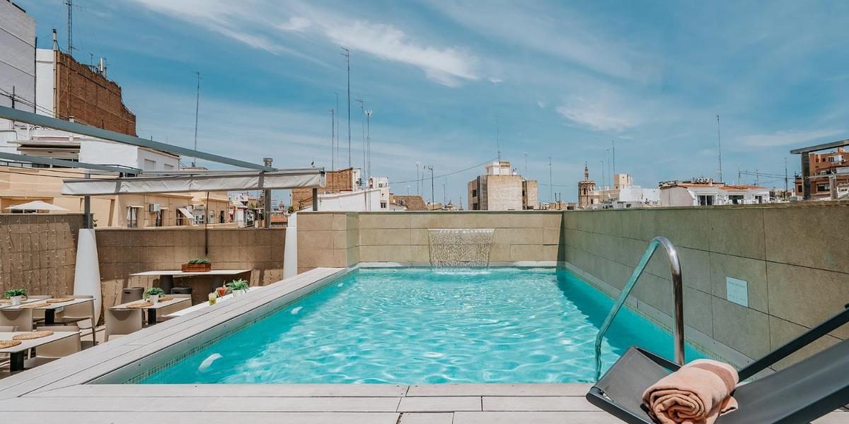 Rooftop pool with a metal ladder, rolled towels and surrounding city buildings.