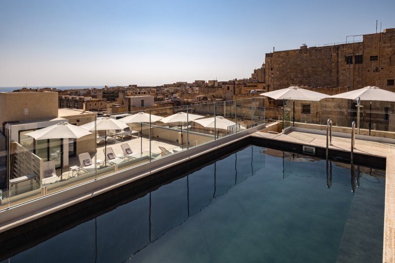 Rooftop pool beside a glass railing and Valletta stone buildings at Palais Le Brun.