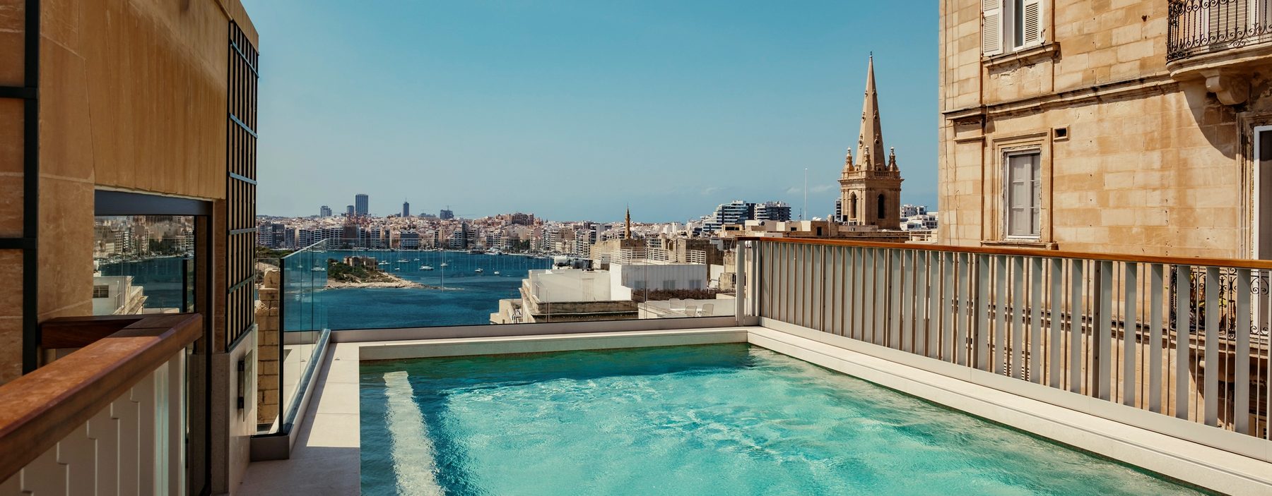 Rooftop pool beside Valletta stone buildings with harbour in the distance.