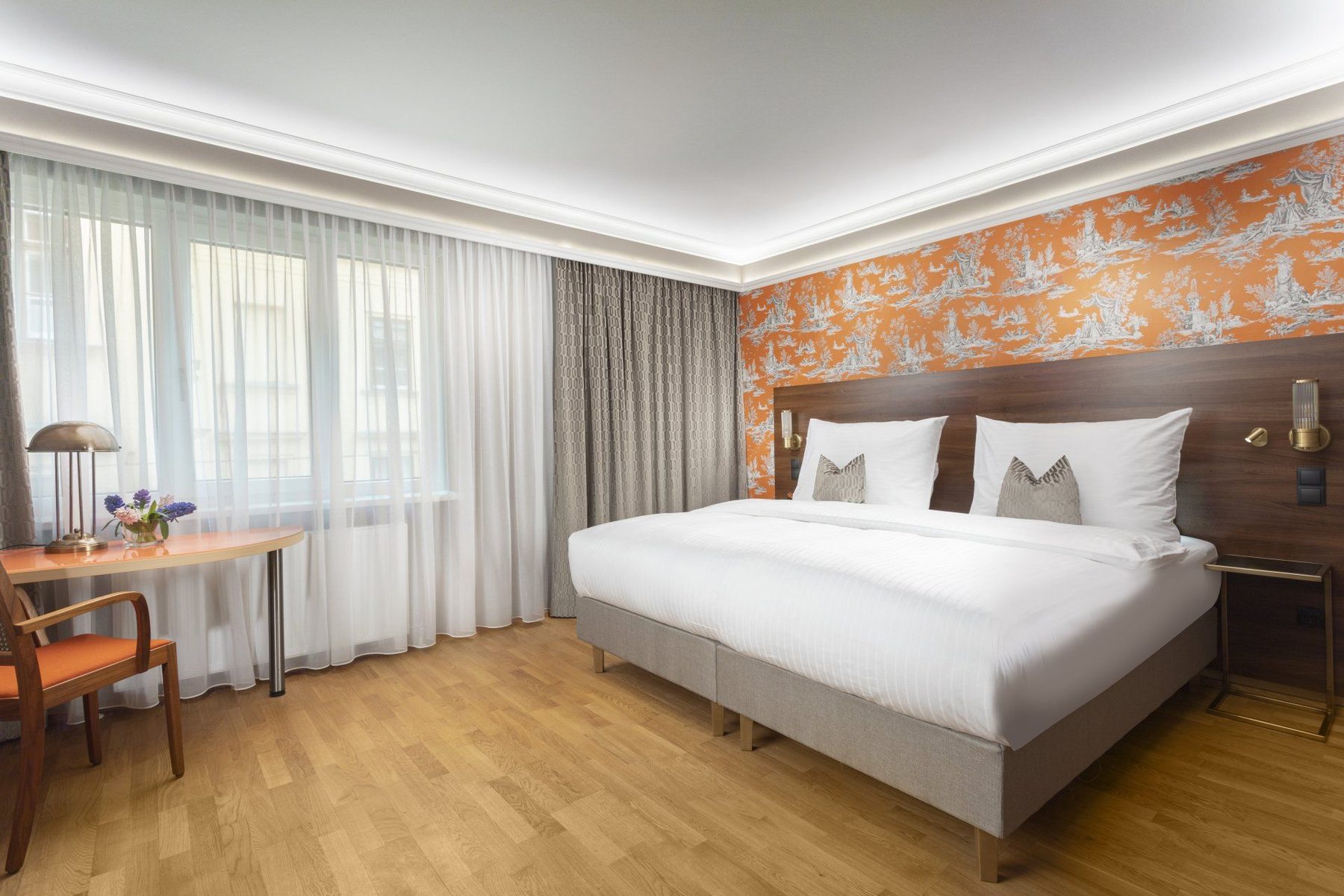Bed against orange patterned wallpaper with a desk beside the window.