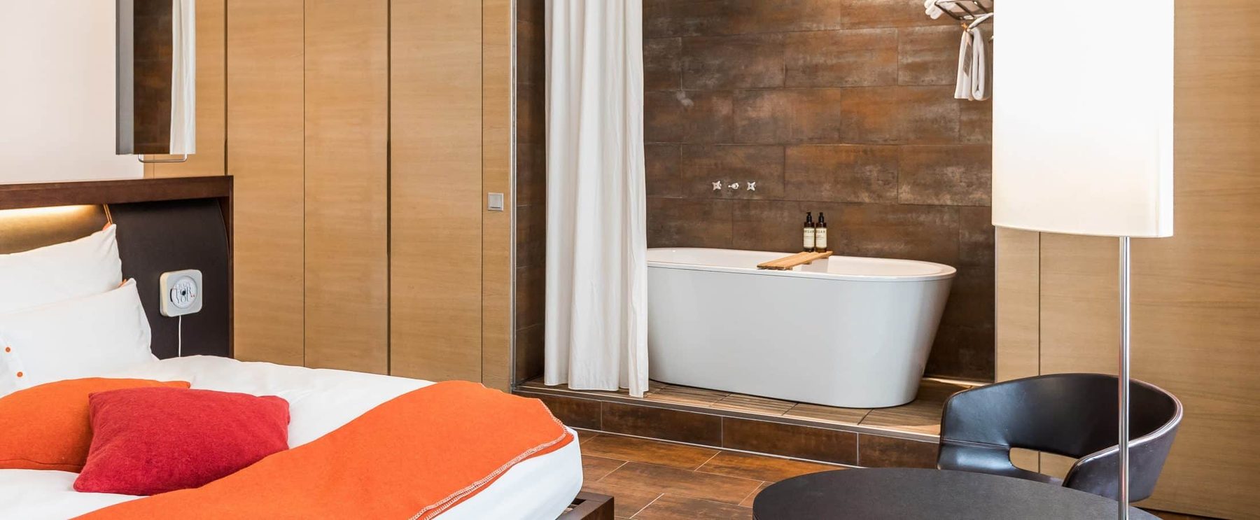 Bed with orange throw and freestanding bathtub behind a curtain in a Beletage XL room.
