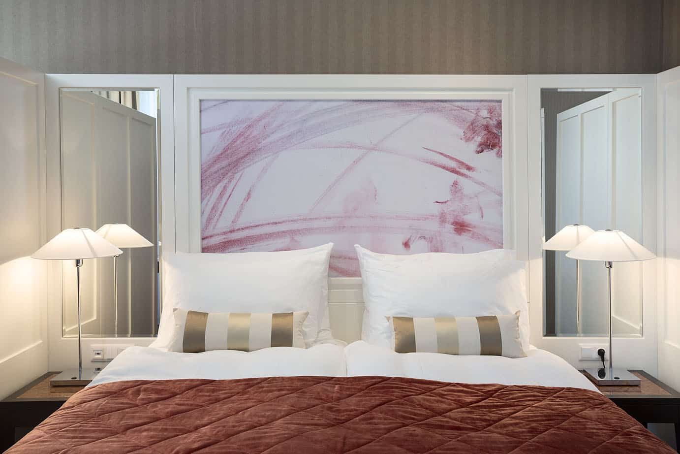 Bed with a red cover beneath a pink abstract artwork at The Harmonie Vienna.