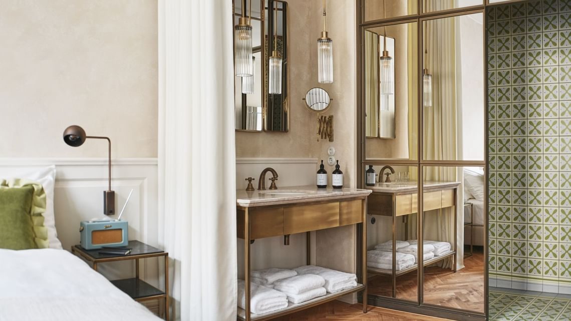 Glass and brass-framed bathroom enclosure beside a bed in a MOTTO DELUXE room.